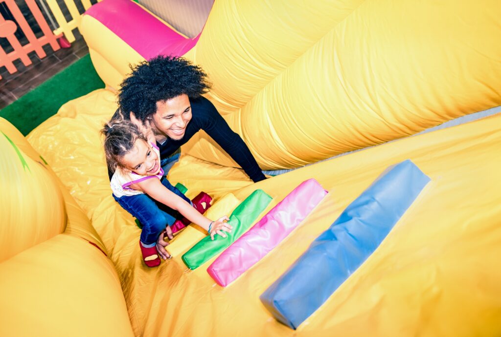 Latin american dad playing with mixed race daughter on inflatable slide at kindergarten playroom